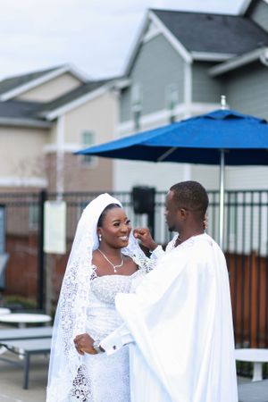 Newlywed couple in traditional white wedding attire share a tender moment by a blue umbrella.