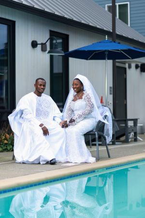 Wedding couple in matching white outfits sit poolside under blue umbrella.