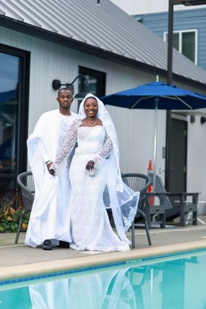 Newlyweds in flowing white garments pose together near poolside with blue umbrella.