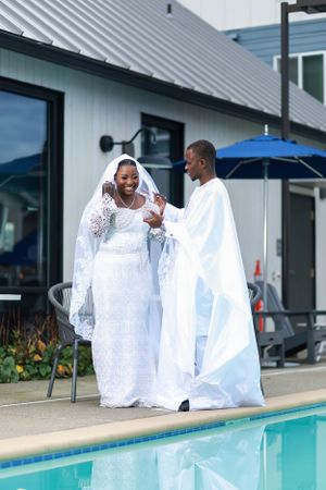 Wedding couple share a laugh while standing by pool in matching white ceremonial dress.