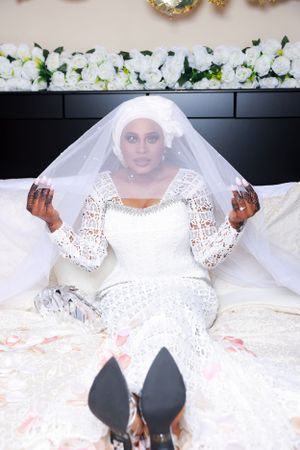 Person wearing white lace wedding dress and veil poses joyfully while lying on white bedding.