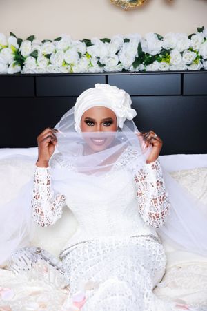 Person wearing elegant white lace gown and veil reclines on white bedding during a photoshoot.