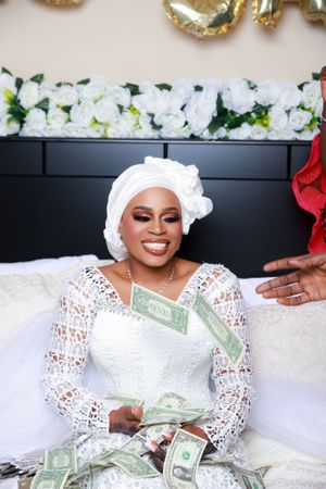 Person in white wedding dress smiles while surrounded by dollar bills on white bedding.