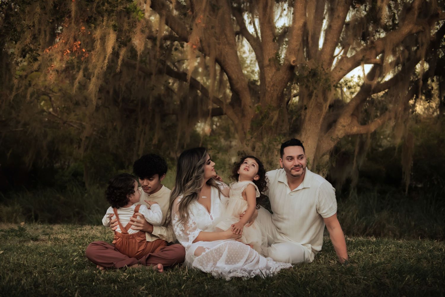 A family sits together on a blanket under a majestic oak tree draped with Spanish moss at sunset.