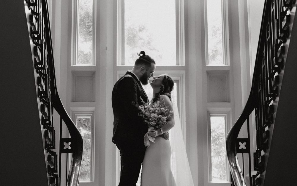 A bride and groom in an intimate moment on an elegant staircase with tall windows, captured by Apertured Media Auckland Wedding Photographer