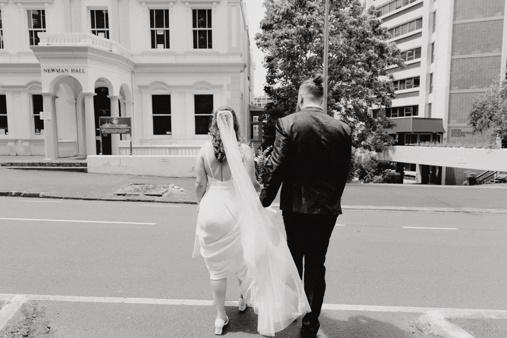 A couple in wedding attire walking down a city street, captured by Apertured Media Auckland Wedding Photographer.