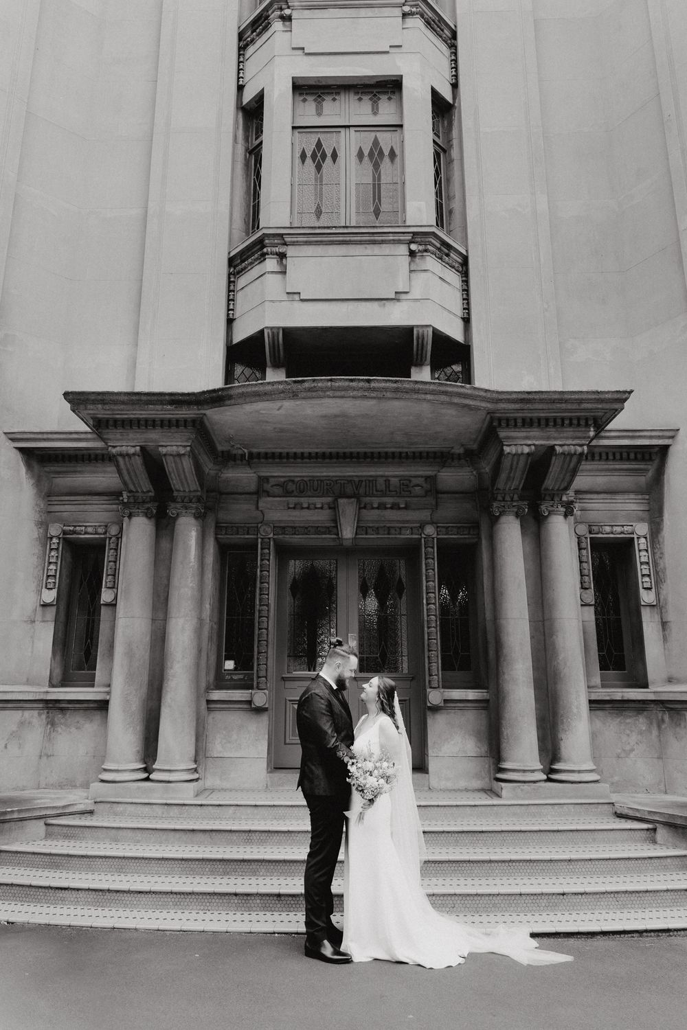 A couple in formal wedding attire stands on grand stone steps, captured by Apertured Media Auckland Wedding Photographer.
