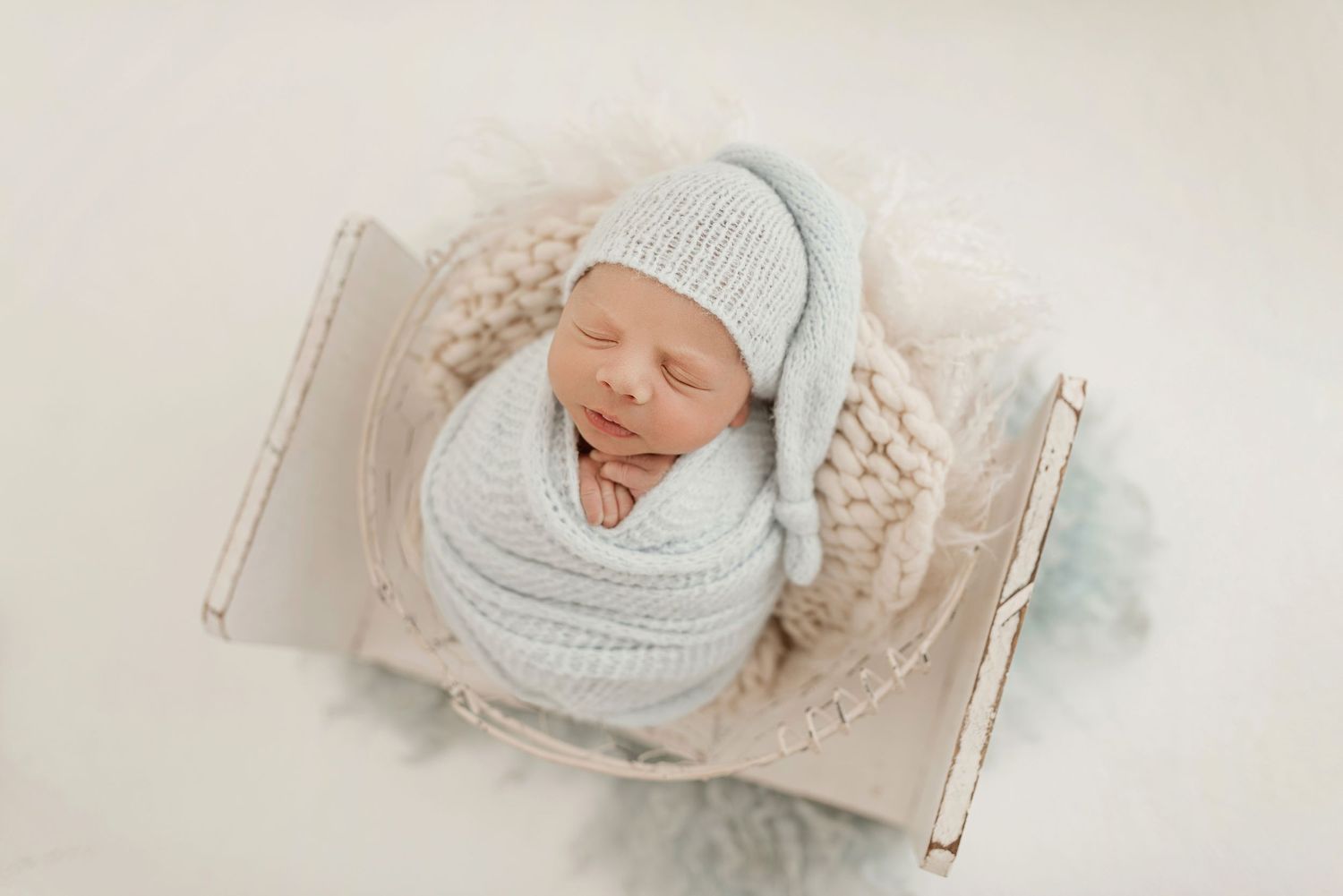 Newborn baby wearing light blue knit hat and wrap sleeps peacefully in soft cream blanket.