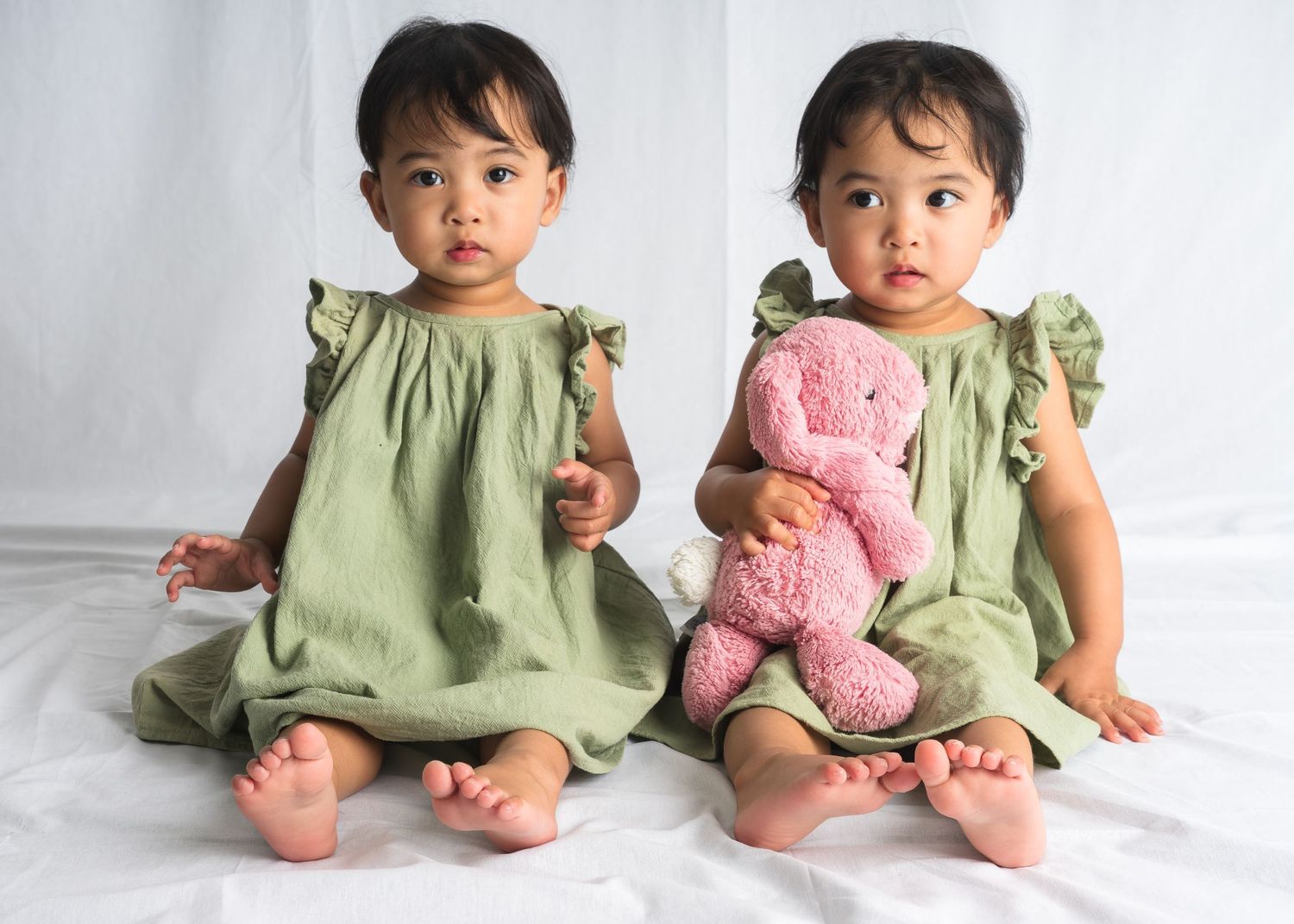 Two small children in matching sage green dresses sitting with a pink teddy bear.