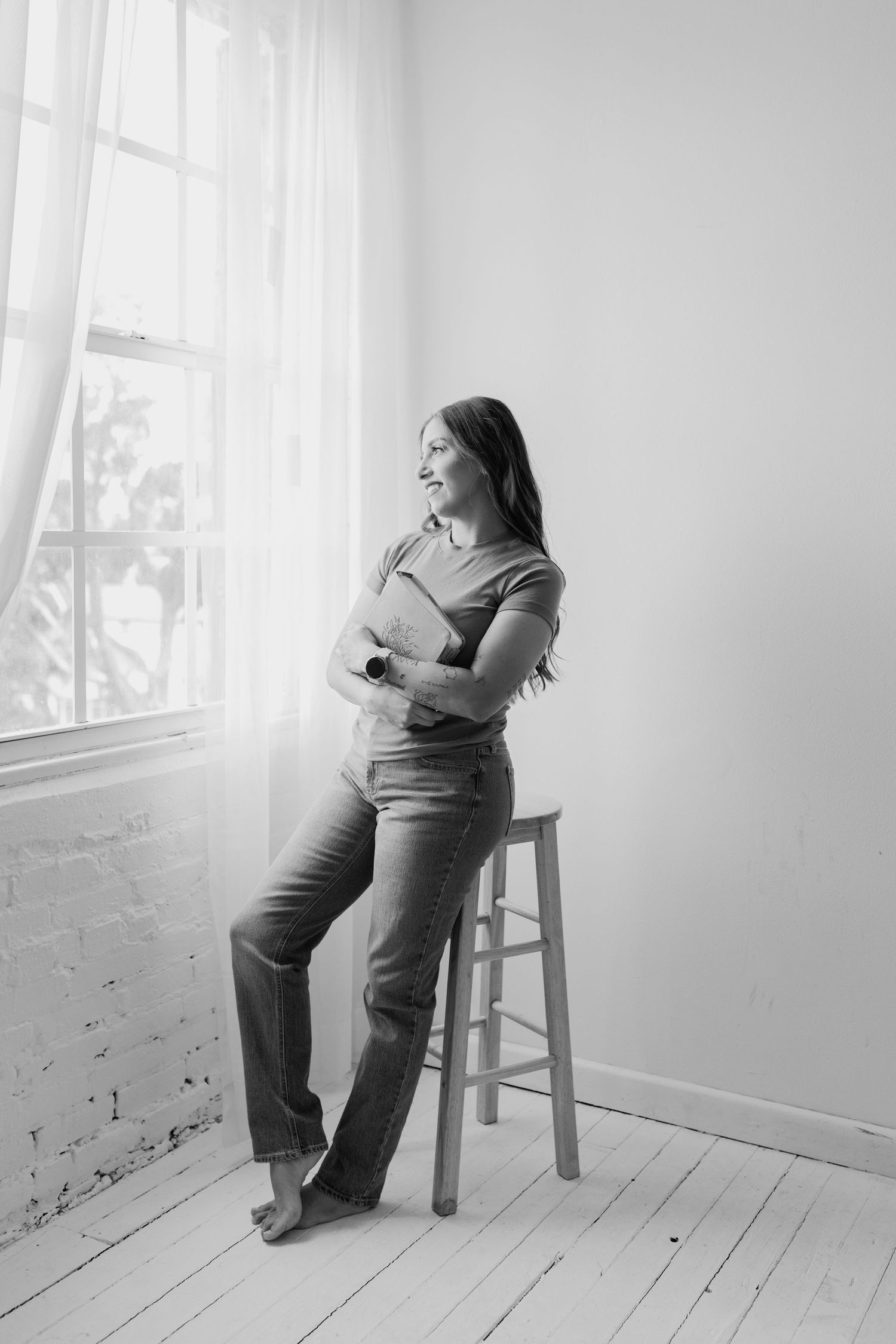 Black and white branding portrait of a photographer sitting on a stool near a window.