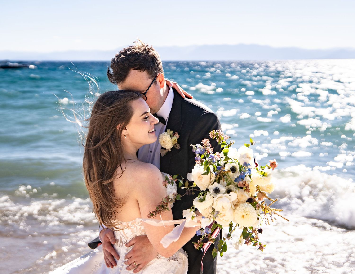 A loving couple embraces on a sunny beach near Hyatt holding a bouquet of white flowers with sparkling Lake Tahoe waves behind them.