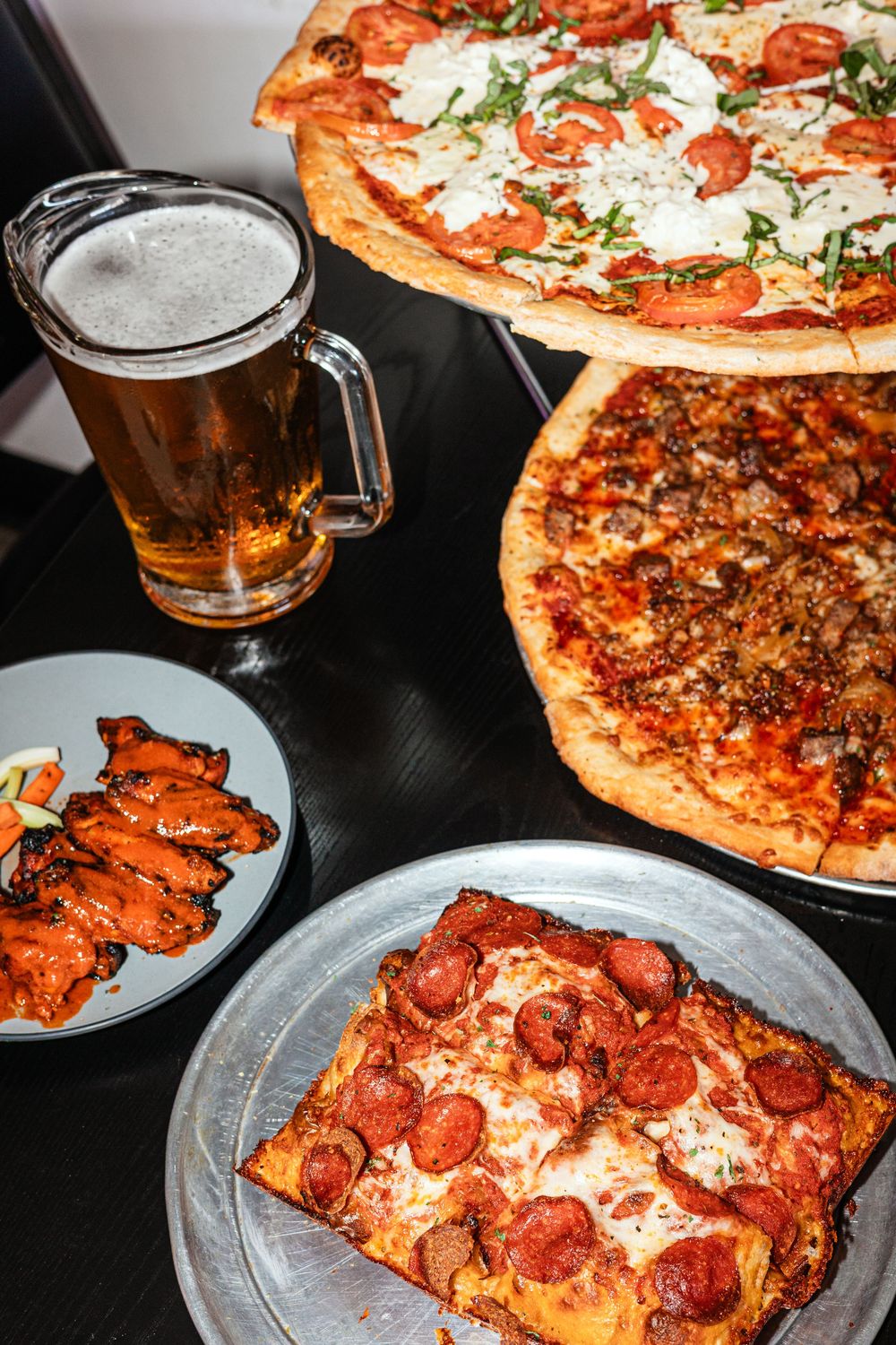 A spread of two pizzas, a square pepperoni slice, chicken wings, and a cold beer mug on a dark table.