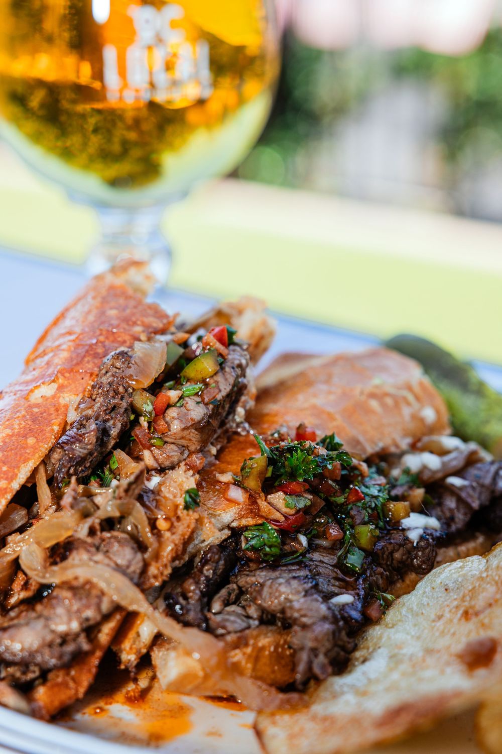 Grilled steak topped with sauteed onions, peppers, and herbs served on a plate with a glass of beer.