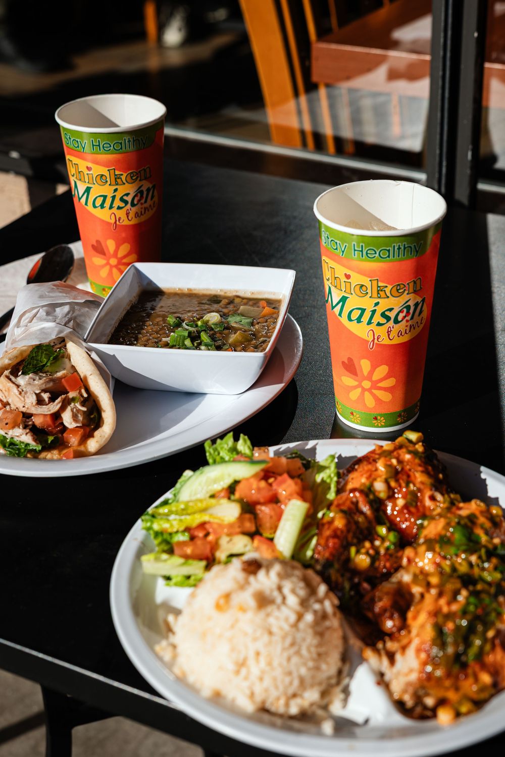 Chicken Maison restaurant meal with rice, glazed chicken, veggies, soup, and branded cups on a black table.