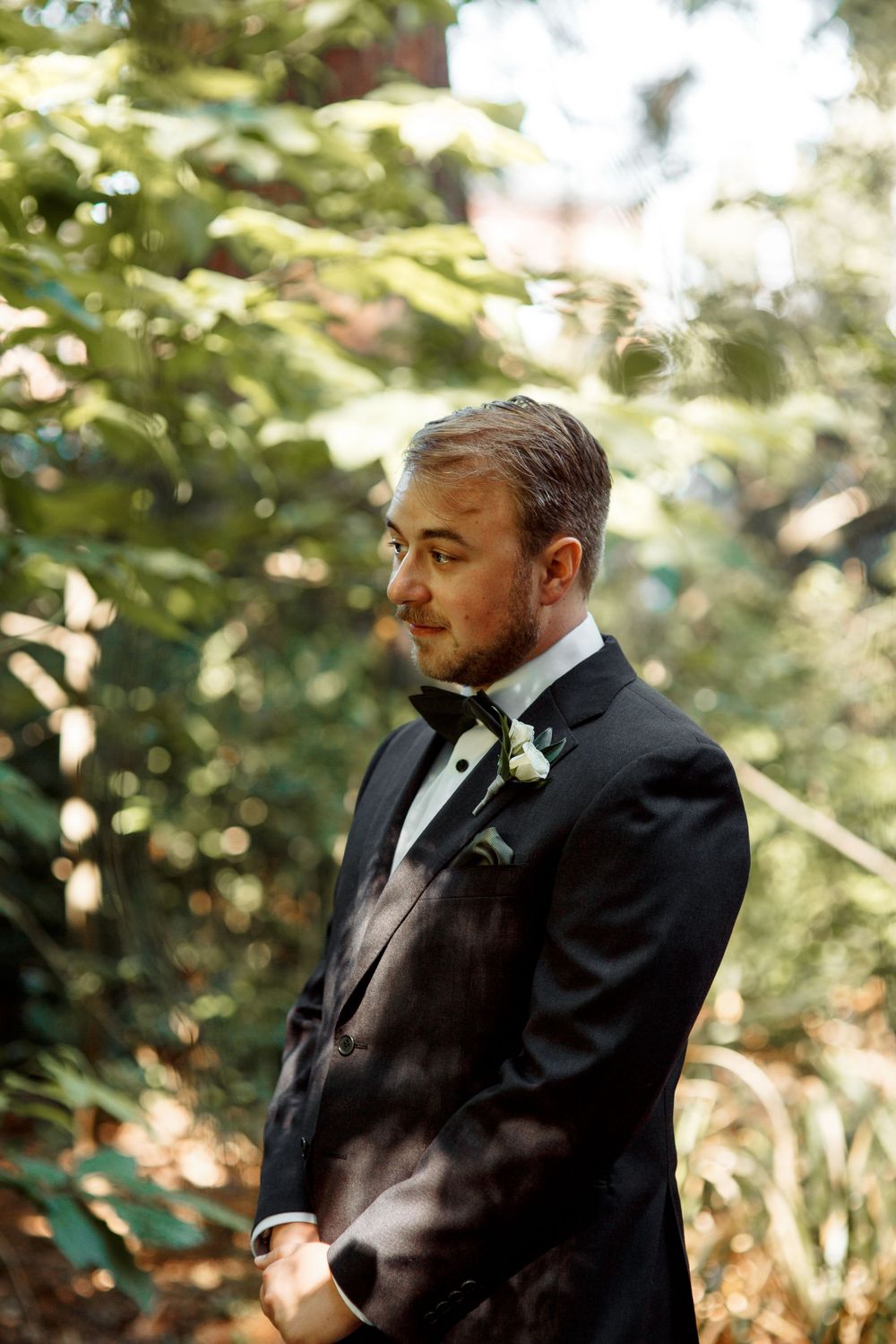 Groom in classic black tuxedo with boutonniere stands in dappled sunlight.