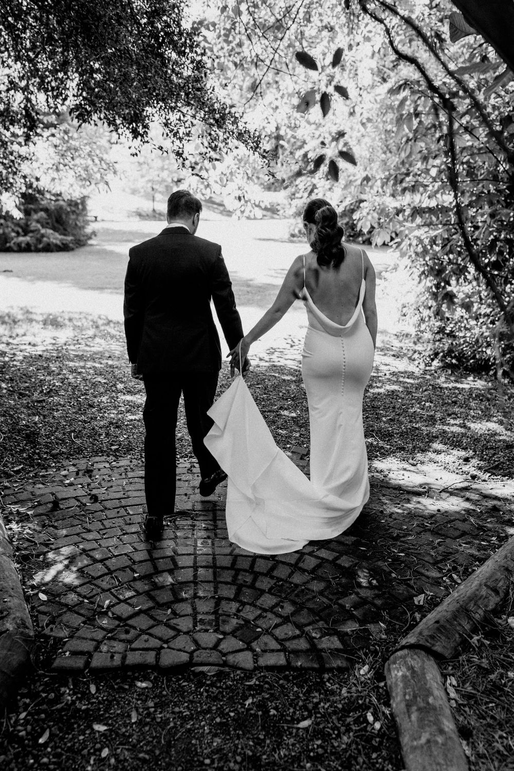 Black and white photo of couple walking hand in hand on garden path after wedding ceremony.