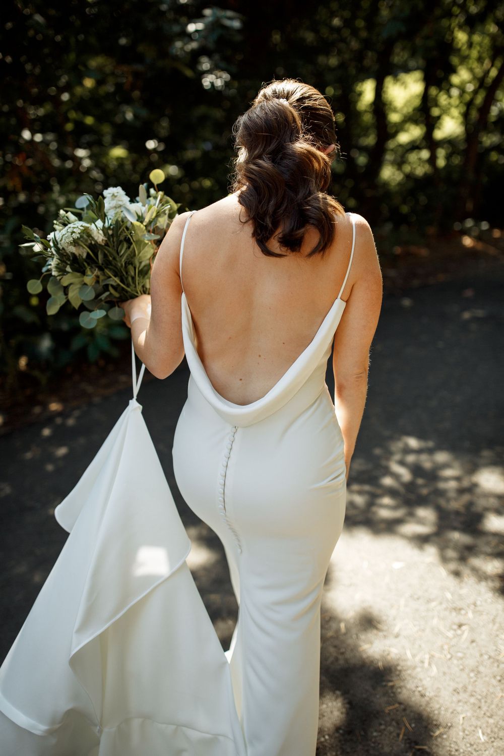 Bride in backless white gown holding a bouquet of eucalyptus on a sunlit garden path.