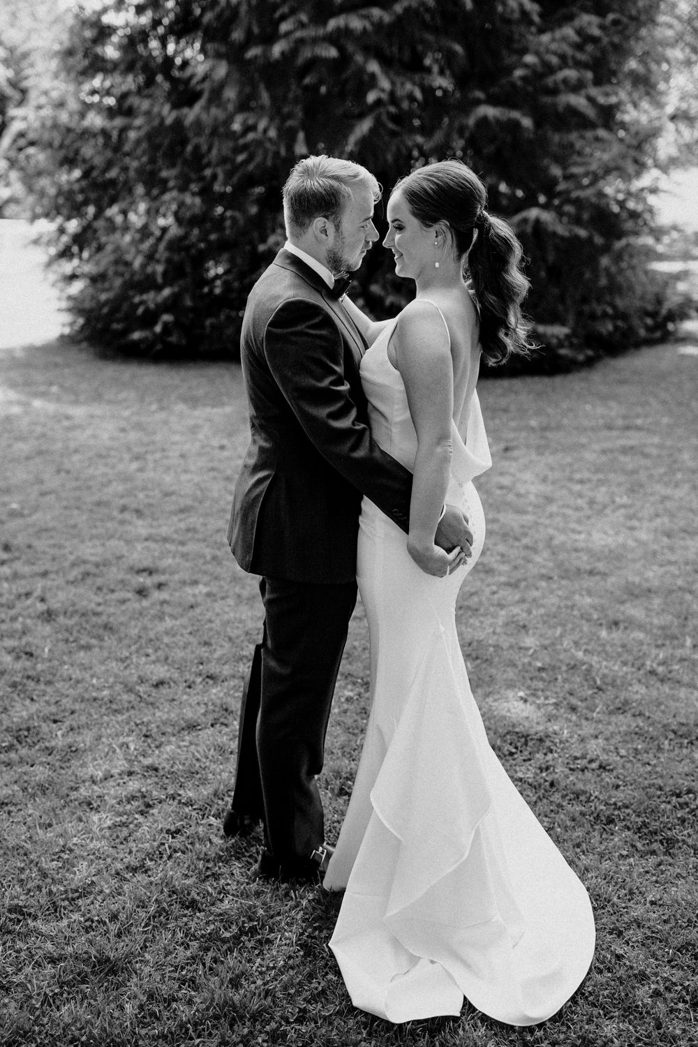 Black and white portrait of couple in elegant wedding attire standing close on lawn.