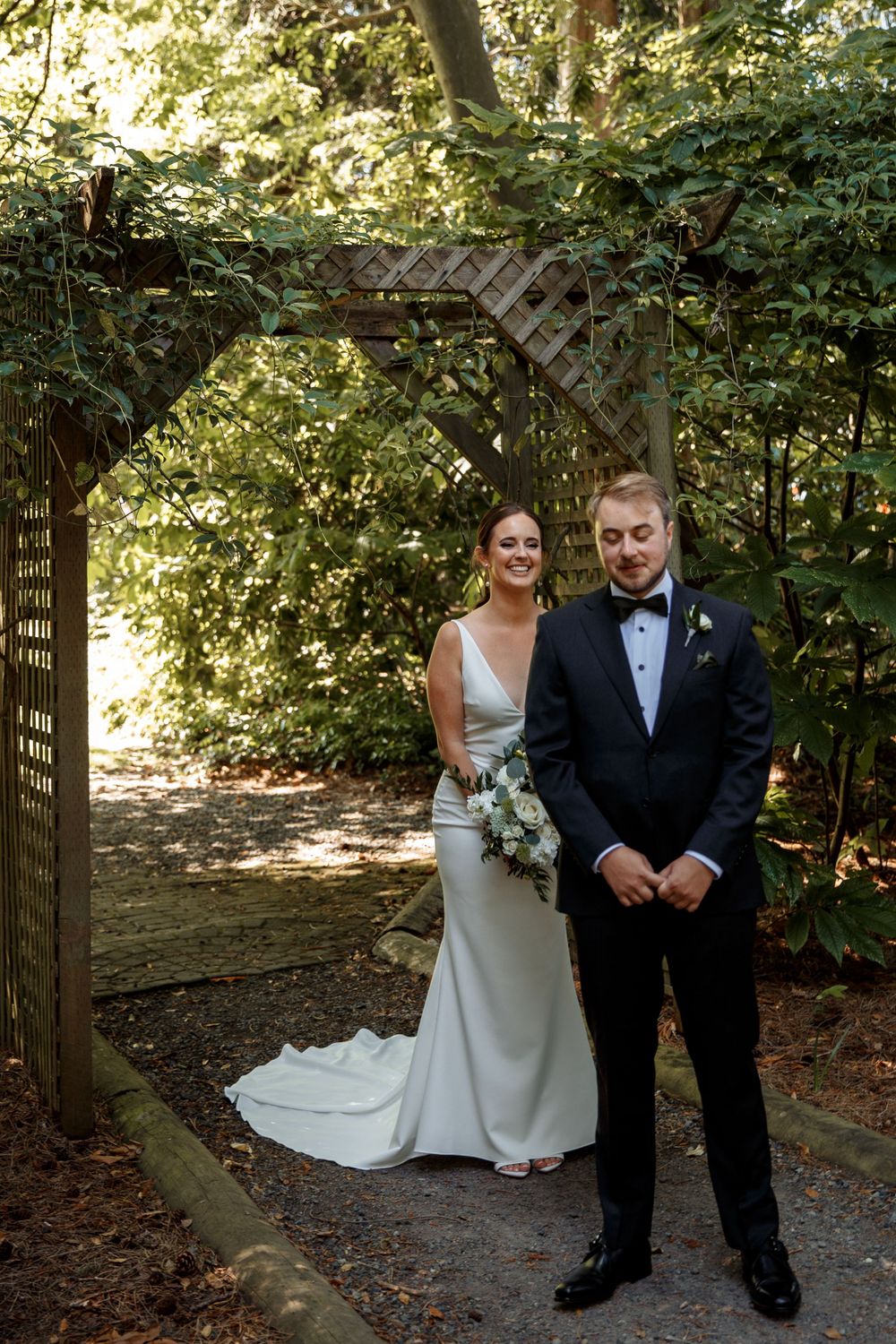Elegant wedding couple posing under a wooden garden arch surrounded by lush greenery.
