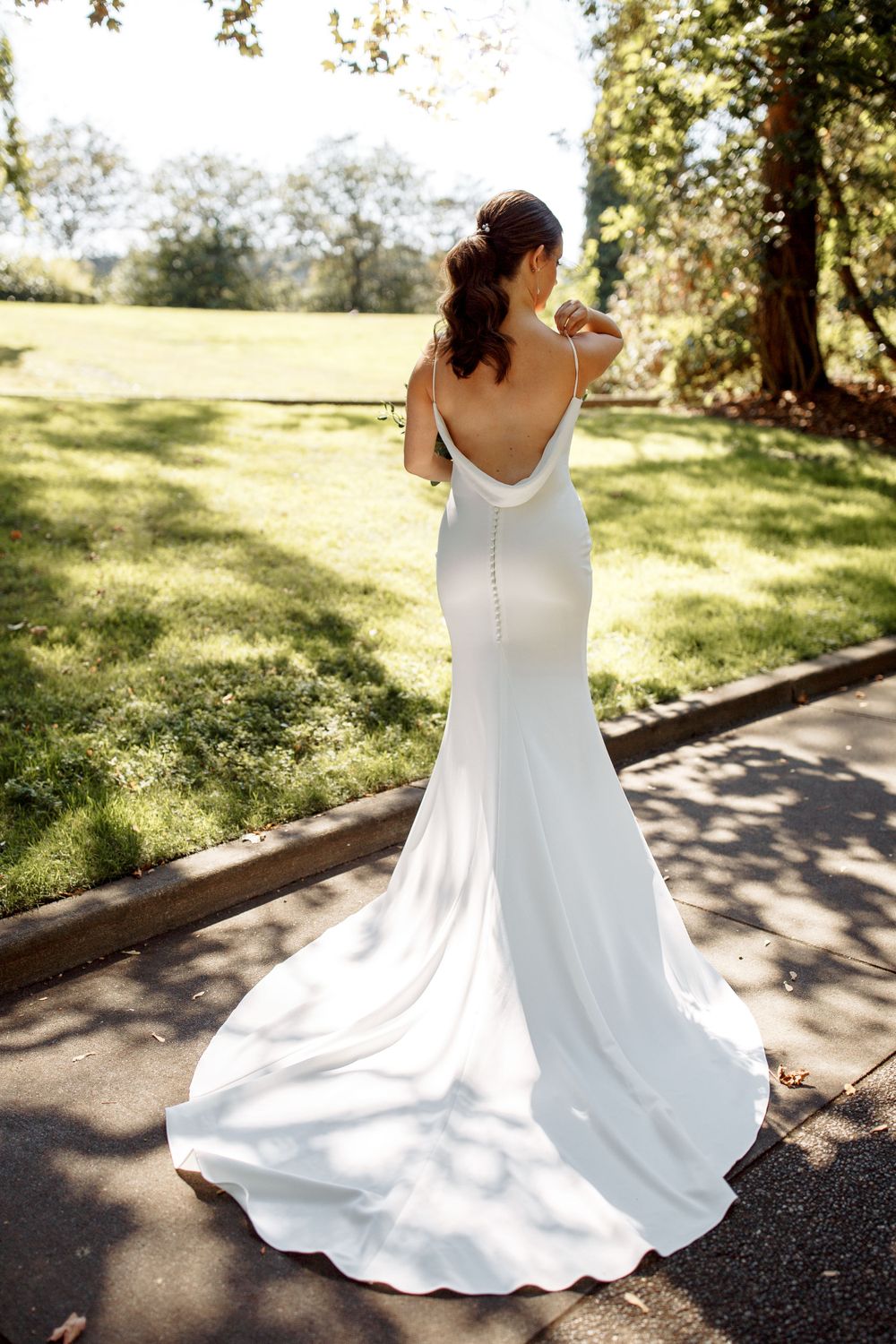 Back view of bride in fitted white wedding dress with train standing in sunlit garden.