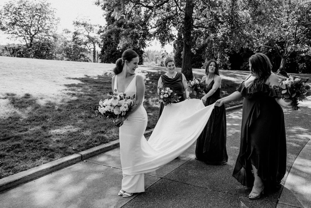 Black and white candid of wedding party helping bride with dress outdoors.