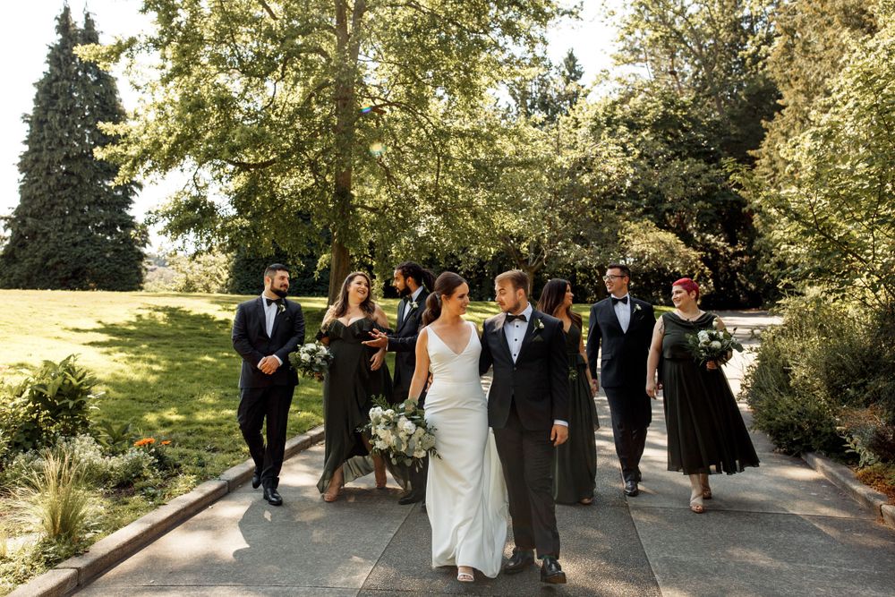Wedding party walks together along an outdoor garden path on a sunny afternoon with trees and greenery in background.