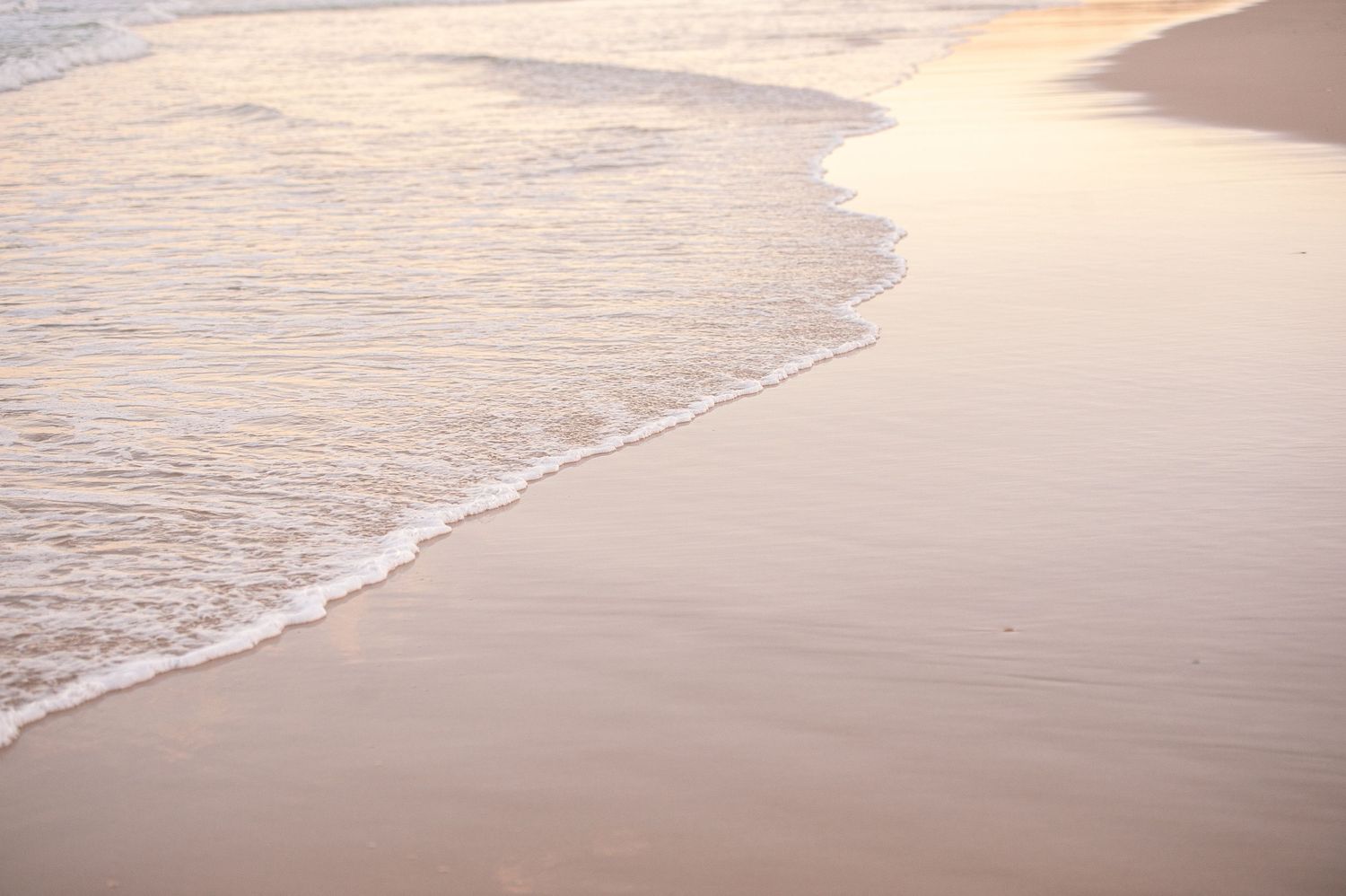 Gentle ocean waves wash up on smooth beige sand during sunrise creating a peaceful beach scene.