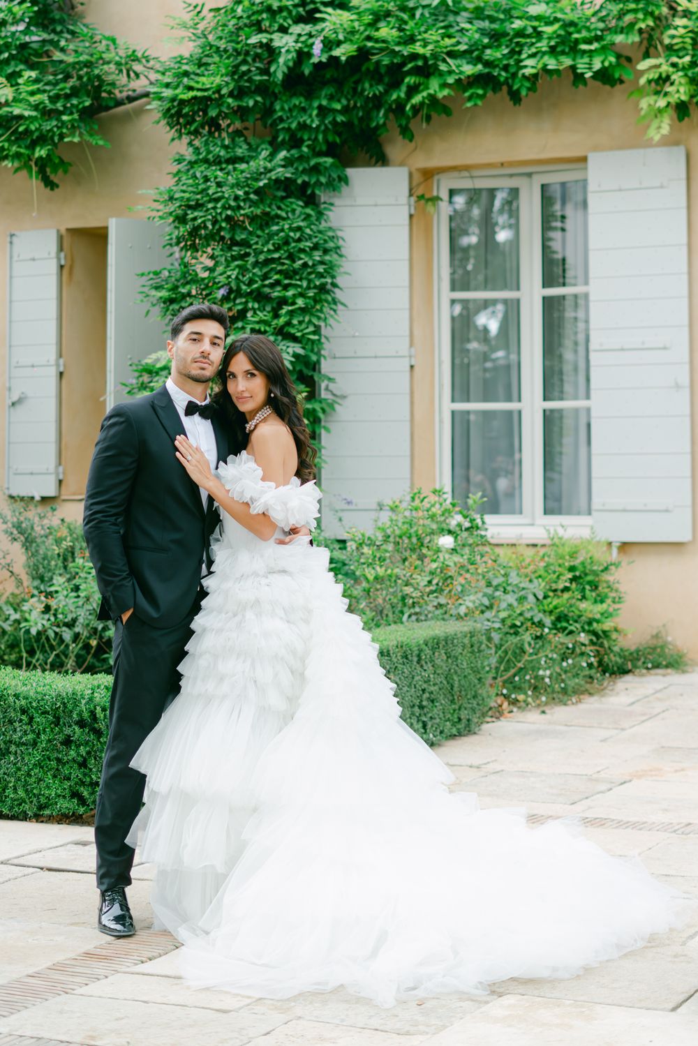 Elegant wedding couple poses in front of a vine-covered building with beige walls and white shutters.