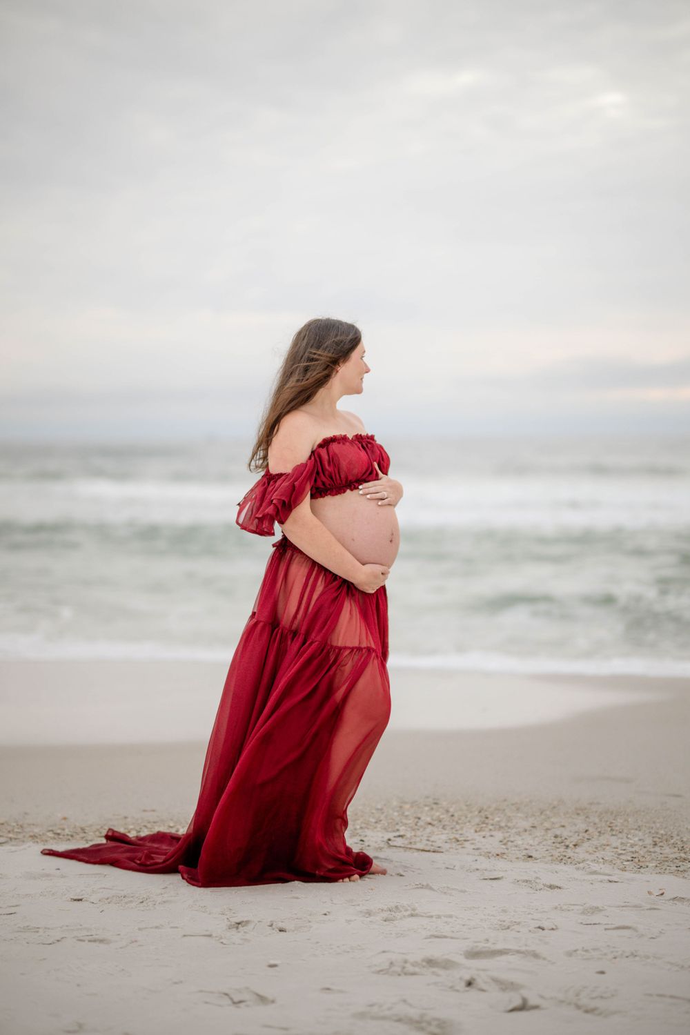 A silhouette of a pregnant woman in a red dress stands against ocean waves.