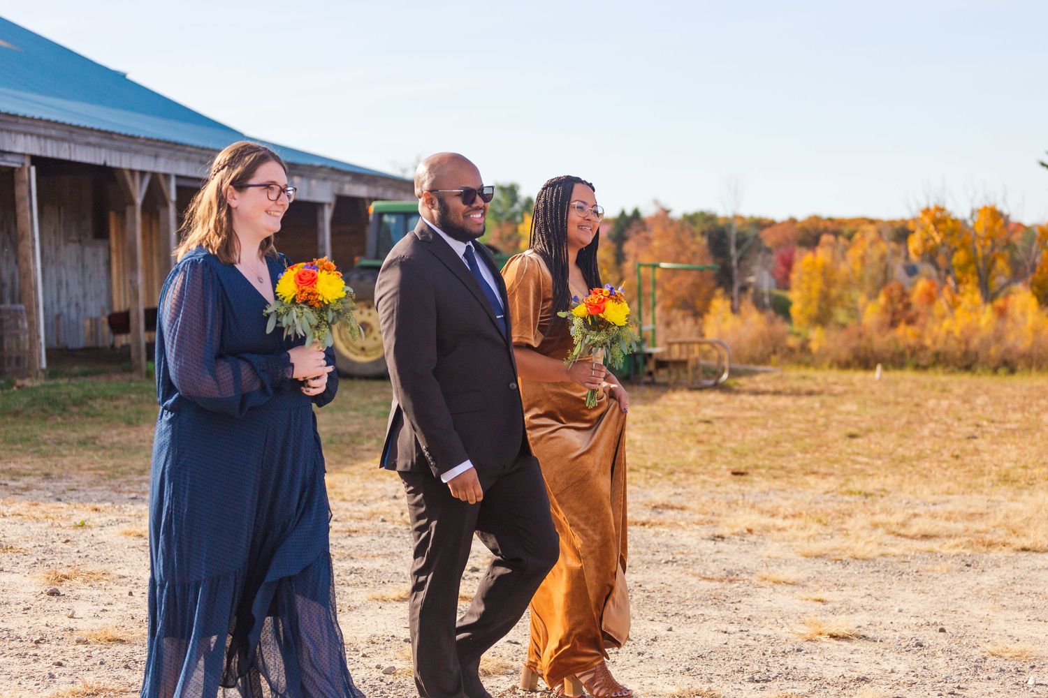 Wedding party holding sunflower bouquets walking outside rustic barn in autumn.