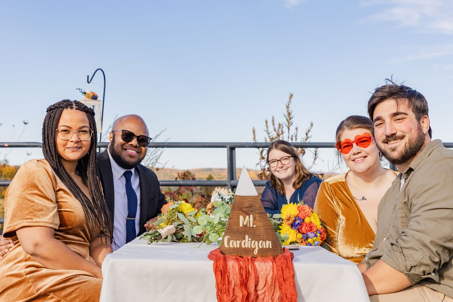 Friends gather for an outdoor celebration at a table decorated with colorful flowers against a scenic autumn backdrop.