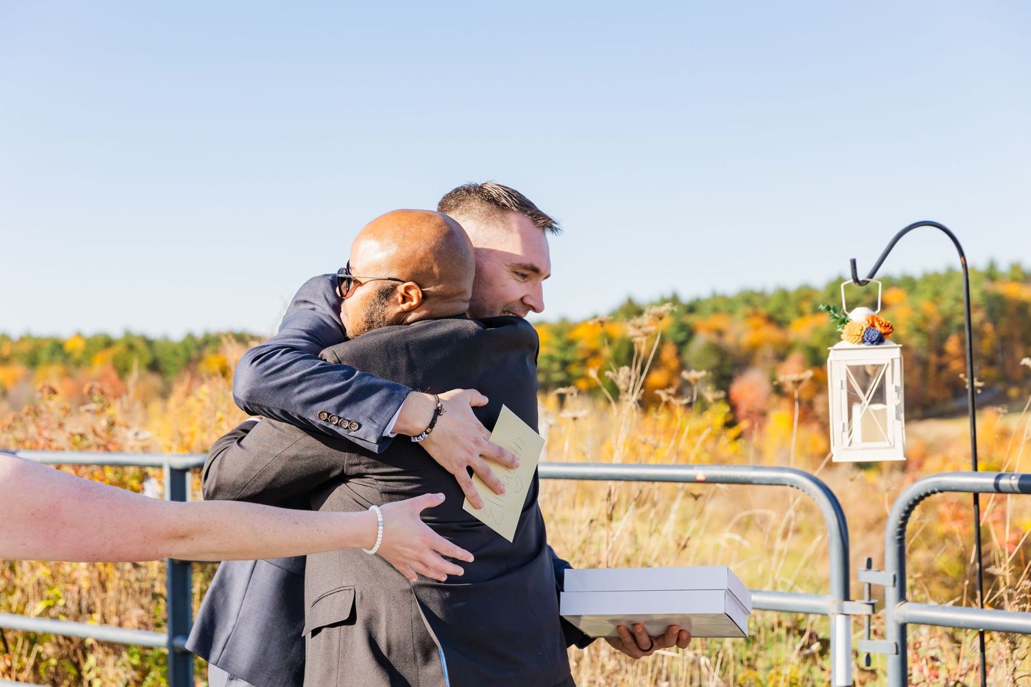 Two people share an emotional embrace on an outdoor deck with fall foliage in the background.