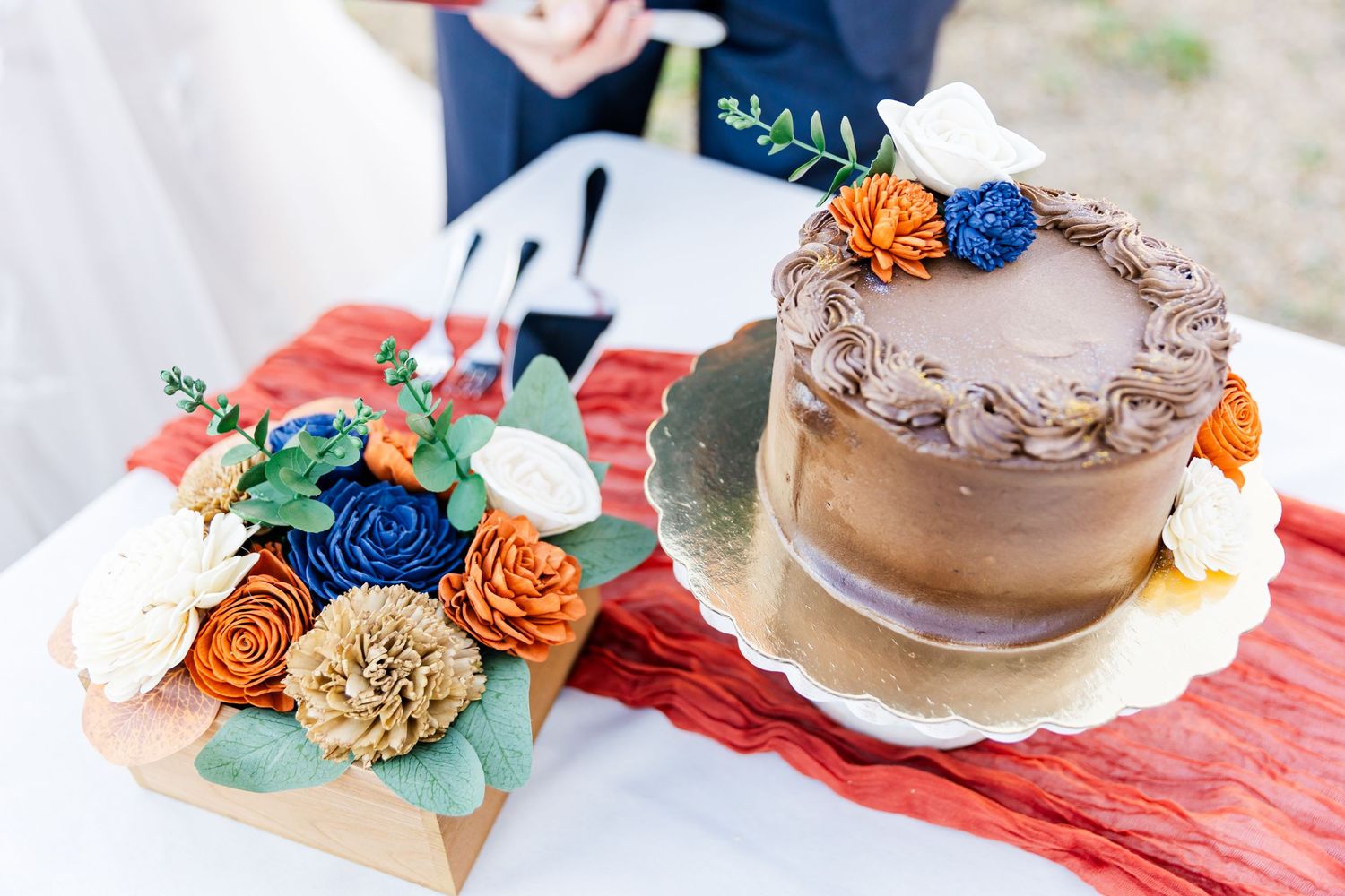 A chocolate wedding cake decorated with orange and blue flowers sits next to a wrapped gift box.