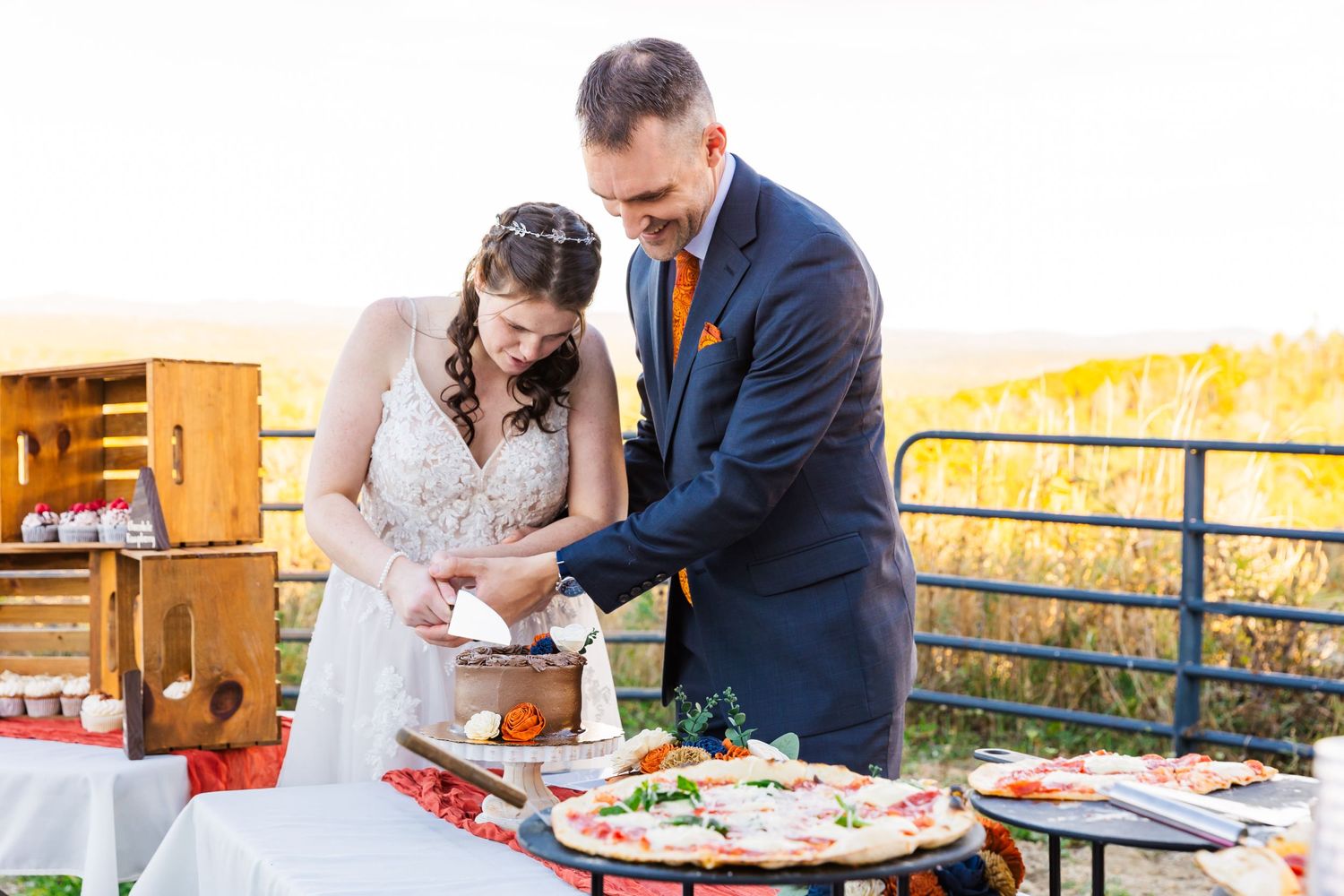 A couple cuts their wedding cake together on an outdoor patio with autumn scenery.