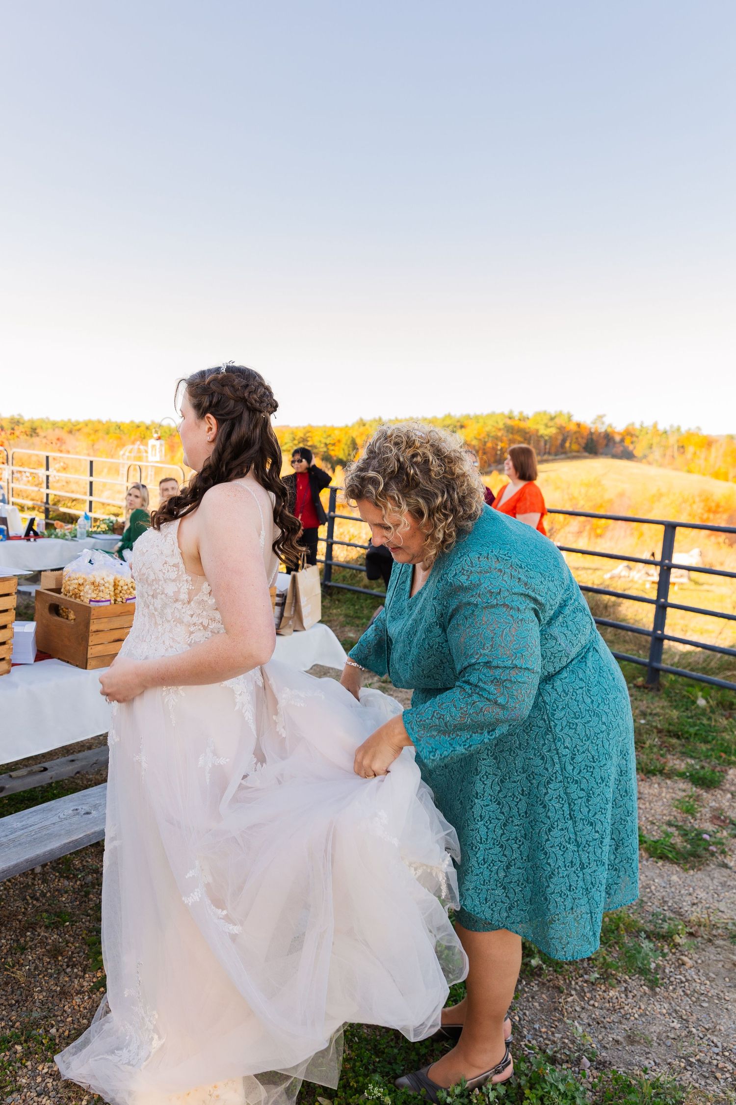 A woman in a teal dress helps adjust the bride's wedding gown outdoors.