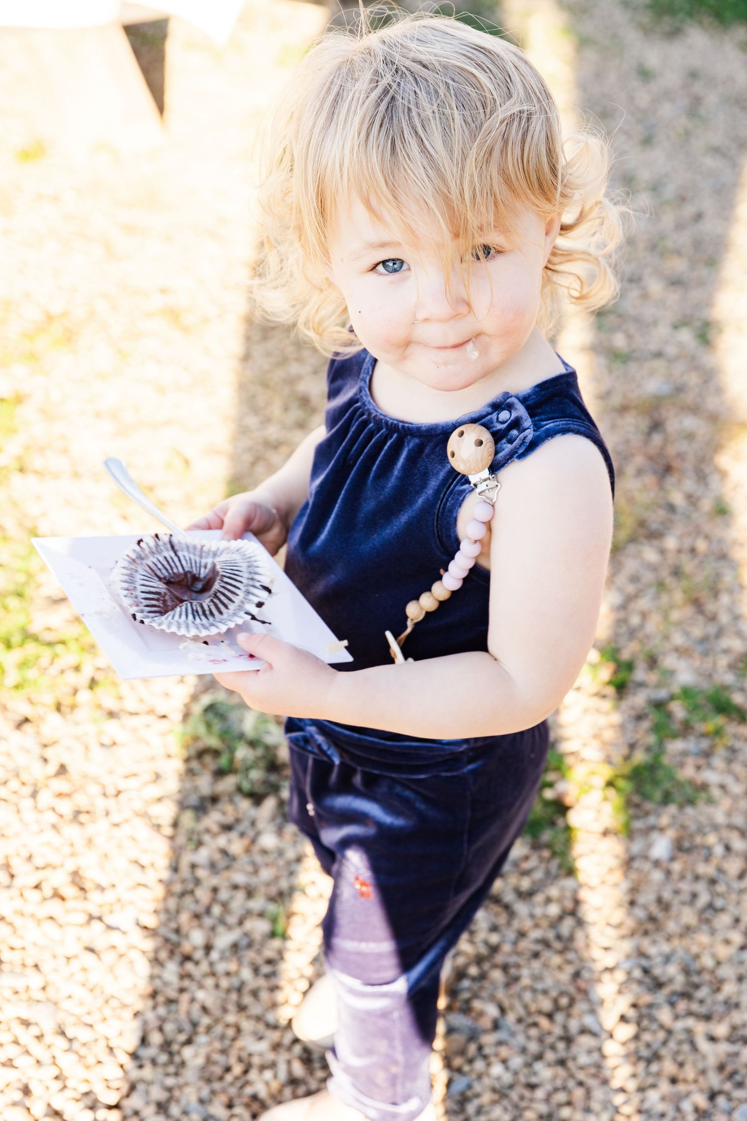 Small child wearing a navy blue dress holds a decorative item while standing in sunlit outdoor setting.