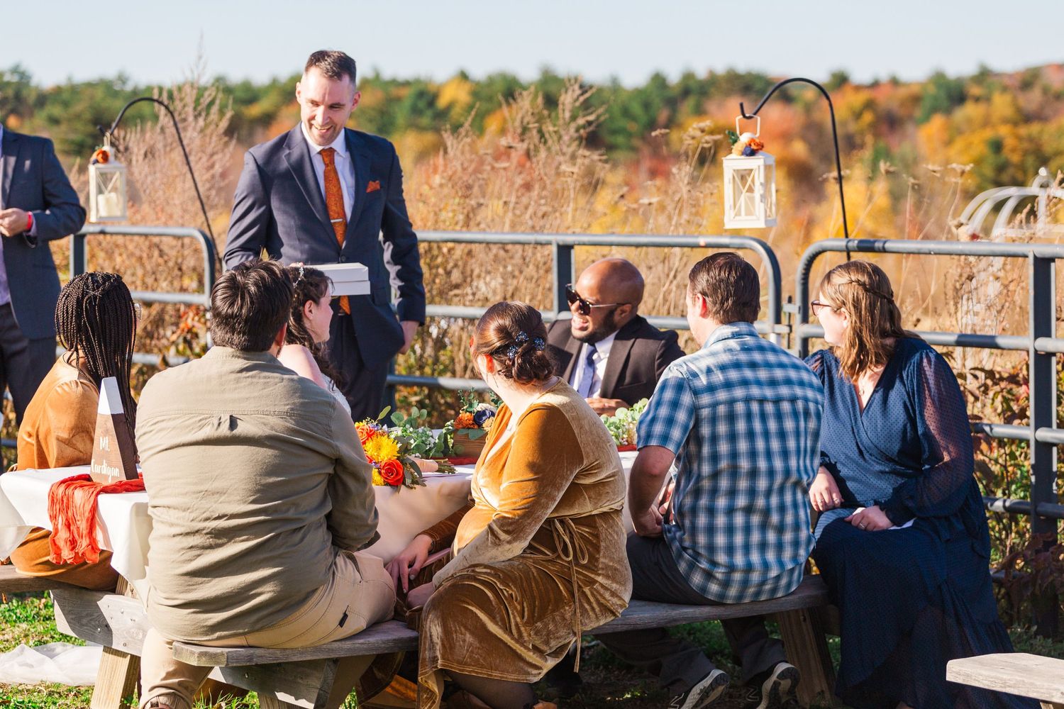 Wedding guests seated at outdoor reception with fall foliage in background.