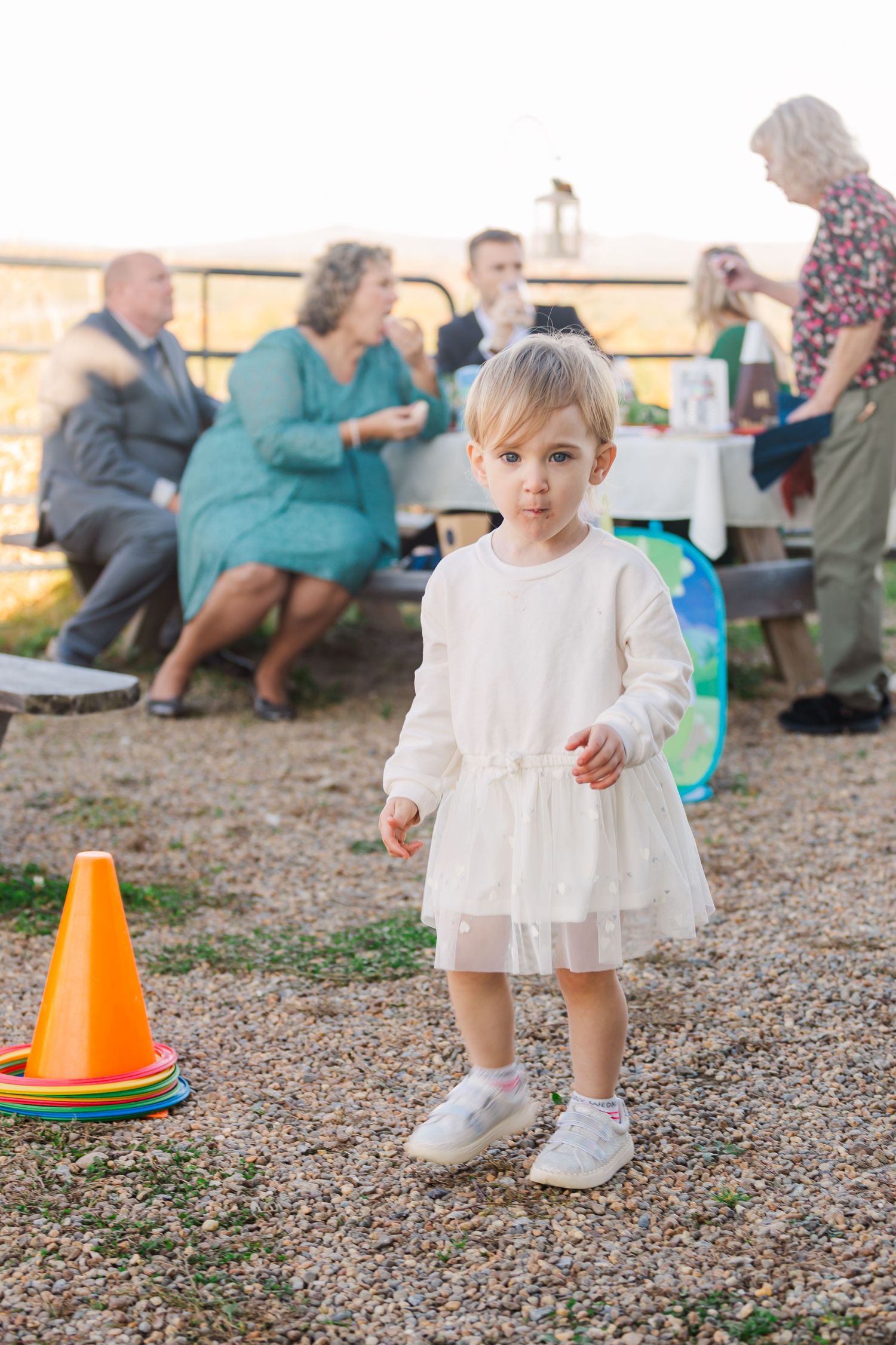 Young child in white dress standing near orange cone at outdoor gathering.