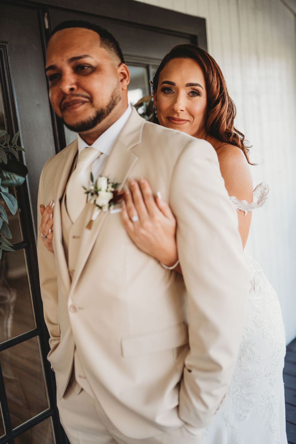 A couple in cream-colored wedding attire embraces in front of a dark doorway.