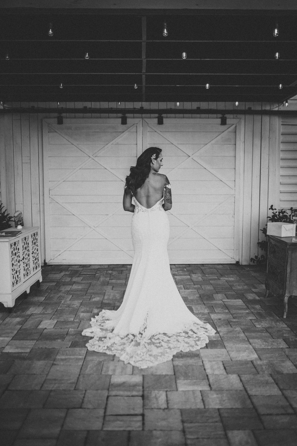 A backlit bride in a fitted white gown with lace train stands against a white barn door.