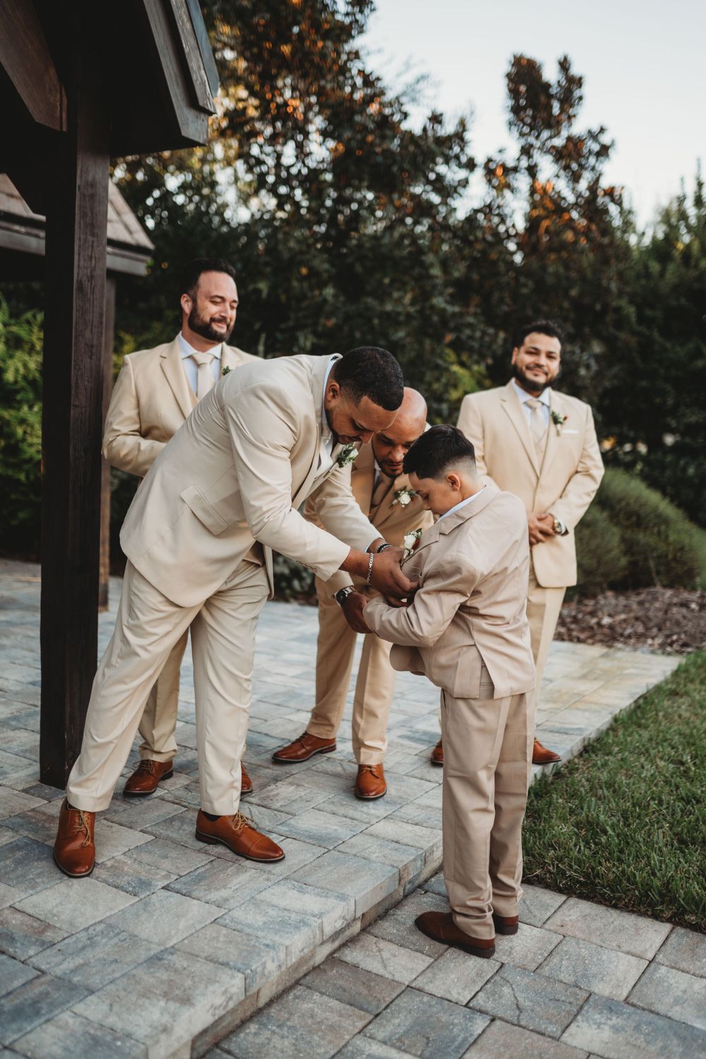 Groomsmen in beige suits share a lighthearted moment with a young ring bearer on stone steps.