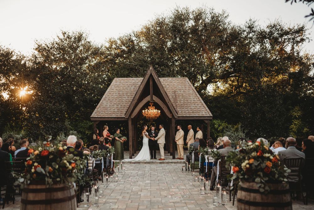 A rustic wooden chapel with floral arrangements hosts an outdoor wedding ceremony at sunset.