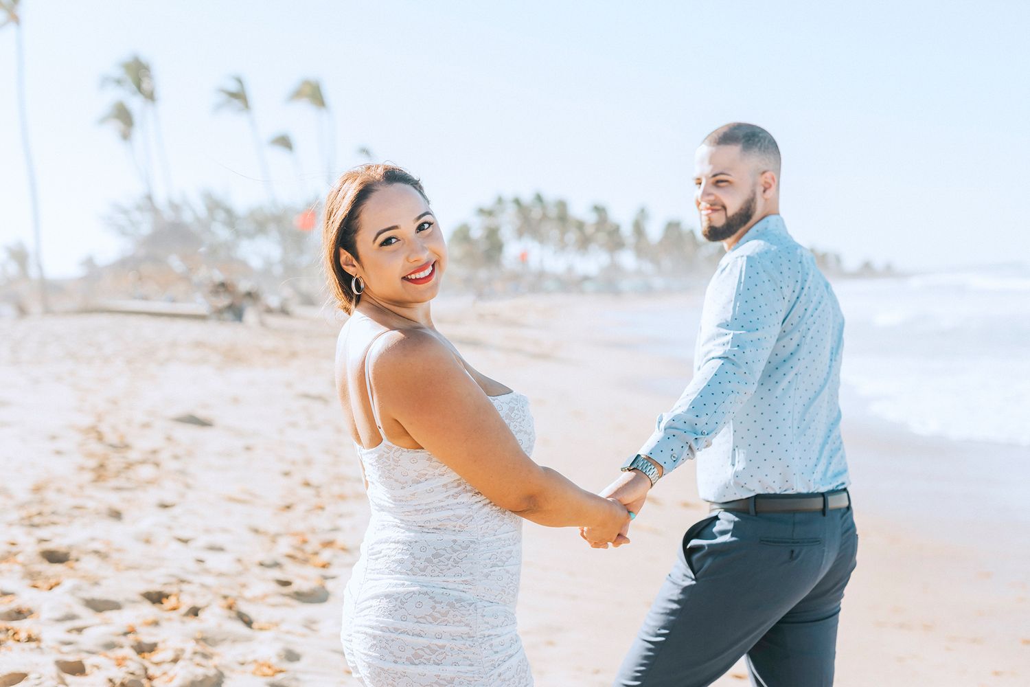 Beach maternity photoshoot with couple in white dress and blue shirt on sandy shore.
