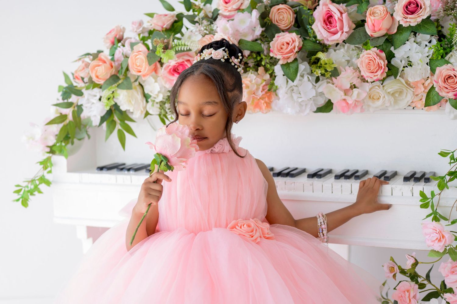 A child in a pink tulle dress poses at a white piano decorated with coral and pink roses white smelling a pink rose.