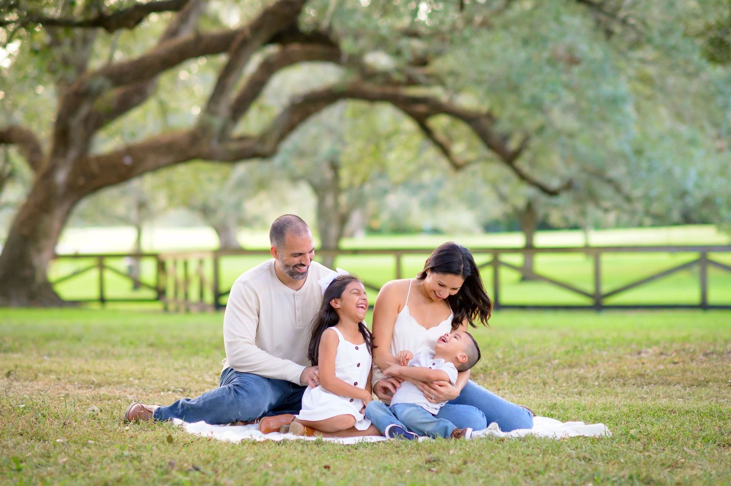 A family sits together on a white blanket under large oak trees in a peaceful park setting.