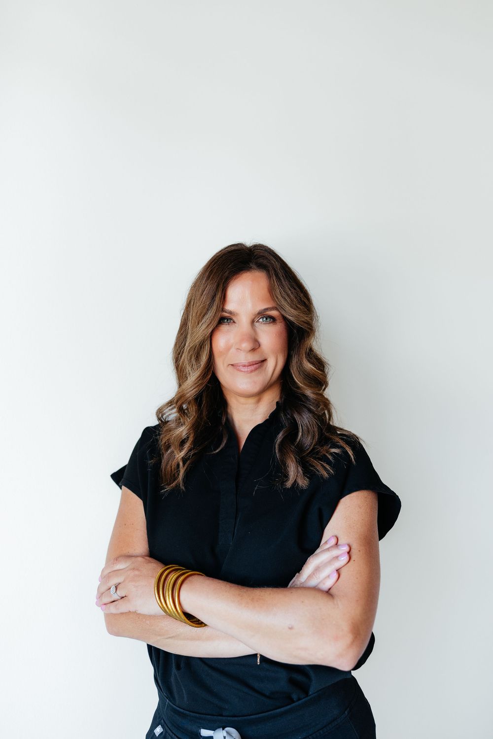 Professional portrait against white wall showing confident pose with crossed arms in black top with styled wavy hair.