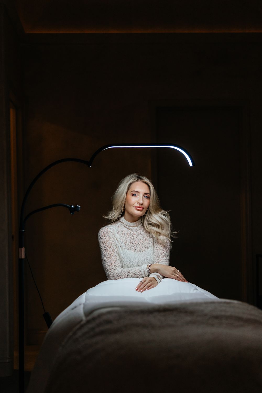 A person in a white lace dress poses on a dark bed in a moody boudoir photoshoot.