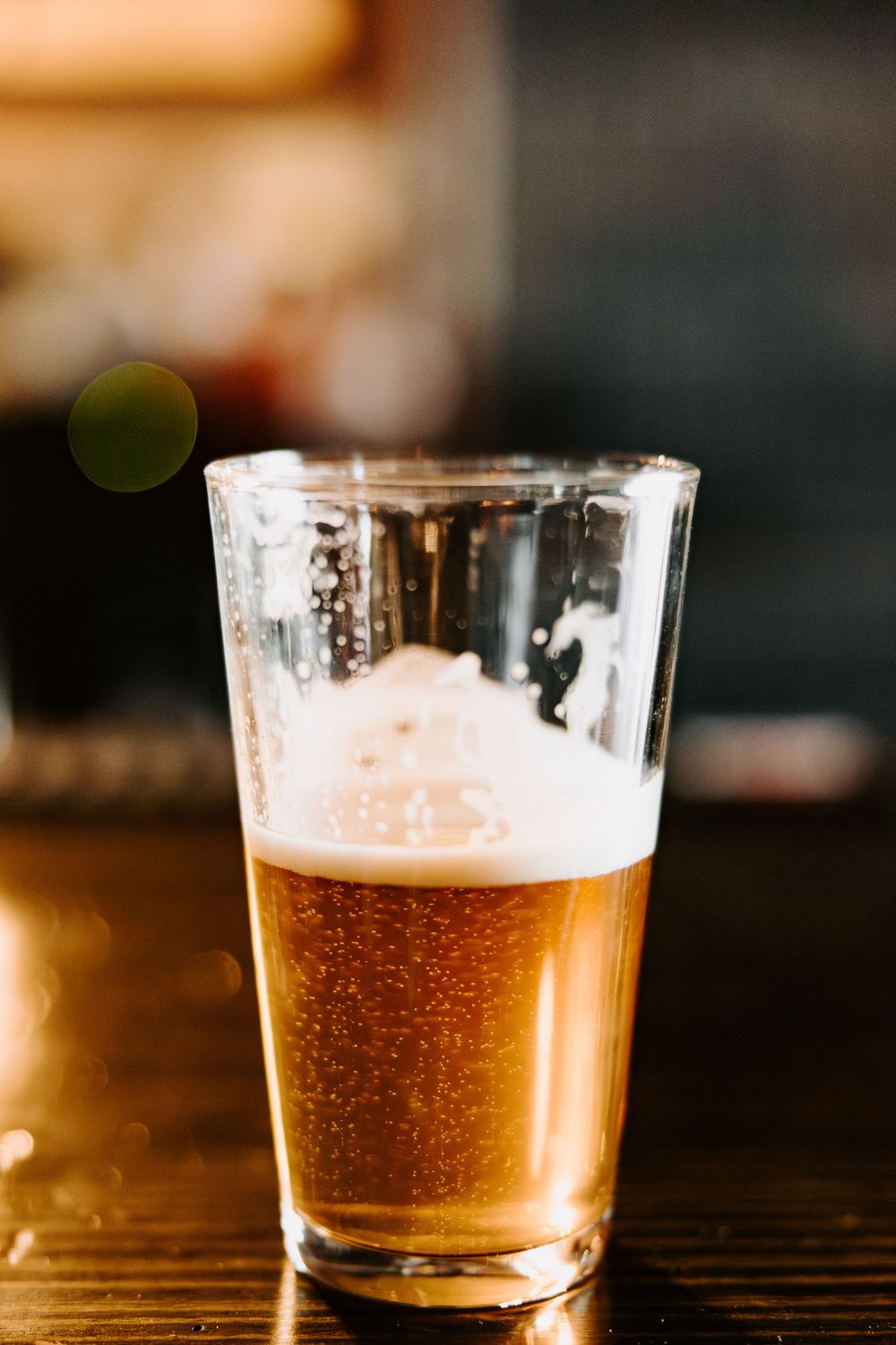 Half-filled glass of amber beer with foam head on dark wooden surface.
