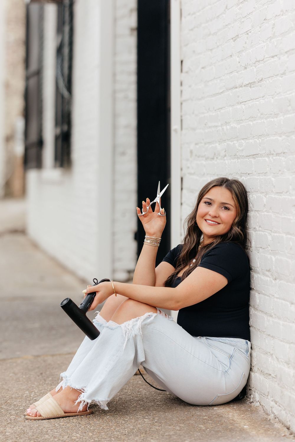 Young woman sitting against white brick wall, smiling, holding scissors and a hair tool in ripped jeans and black top.