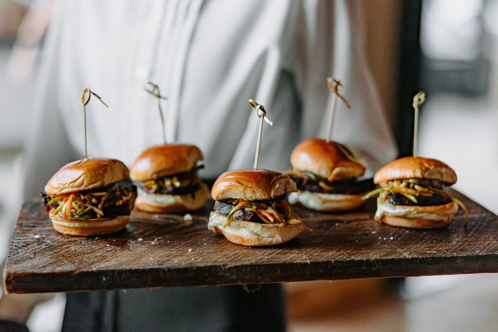 Row of gourmet mini burgers with toothpicks served on rustic wooden board.